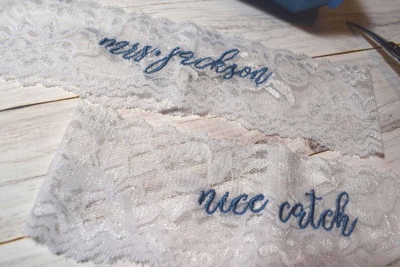 Op de afbeelding: Twee stukken wit kant met blauw geborduurde tekst. Op &eacute;&eacute;n stuk staat "mrs. jackson" en op het andere "nice catch". Het kant ligt op een licht houten oppervlak.