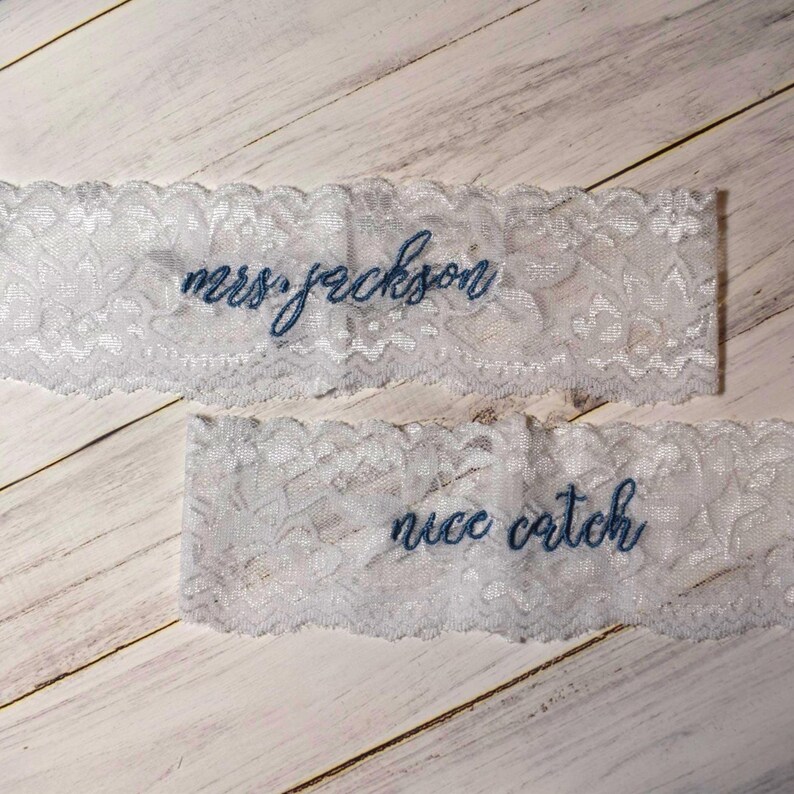 Op de afbeelding: Twee witte kanten kousenbanden met blauw geborduurde tekst. De bovenste kousenband leest "mrs. jackson" en de onderste kousenband leest "nice catch". Het kant heeft een delicaat bloemenpatroon.