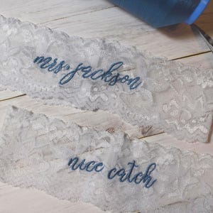 Op de afbeelding: Twee witte kanten kousenbanden met blauw geborduurde tekst. Op &eacute;&eacute;n kousenband staat "mrs jackson" en op de andere "nice catch". Een spoel blauw garen en een schaar zijn ook zichtbaar.