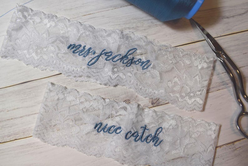 Op de afbeelding: Twee witte kanten kousenbanden met blauw geborduurde tekst. Op &eacute;&eacute;n kousenband staat "mrs jackson" en op de andere "nice catch". Een spoel blauw garen en een schaar zijn ook zichtbaar.