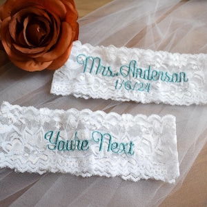 May include: Two white lace garters with teal embroidery. One garter reads "Mrs. Anderson" and "1/6/24". The other garter says "You're Next". A rust-colored rose is in the background.