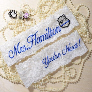 May include: Two white lace garters featuring blue embroidery. One garter displays the text Mrs. Hamilton with a police badge patch numbered 5793. The second garter features the text You're Next! in blue script. The items are surrounded by white pearls.