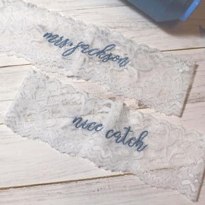 Op de afbeelding: Twee witte kanten kousenbanden met blauw geborduurde tekst. Op &eacute;&eacute;n staat "mrs. jackson" en op de andere "nice catch". Een schaar en een blauw voorwerp staan ook in de afbeelding.