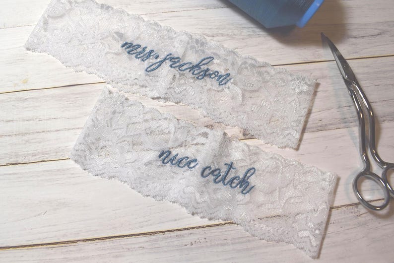 Op de afbeelding: Twee witte kanten kousenbanden met blauw geborduurde tekst. Op &eacute;&eacute;n staat "mrs. jackson" en op de andere "nice catch". Een schaar en een blauw voorwerp staan ook in de afbeelding.