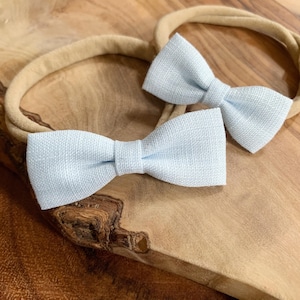 May include: Two light blue bow ties on tan headbands. The bows are made of a textured fabric and are tied in the center. The headbands are made of a soft, stretchy material. The bows are placed on a wooden surface.
