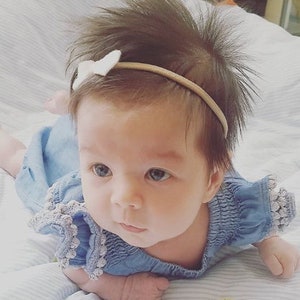 Op de afbeelding: Een babymeisje draagt een lichtblauwe denimjurk met witte bies en een wit strikbandje. Het bandje is een dun elastisch bandje met een klein wit strikje.