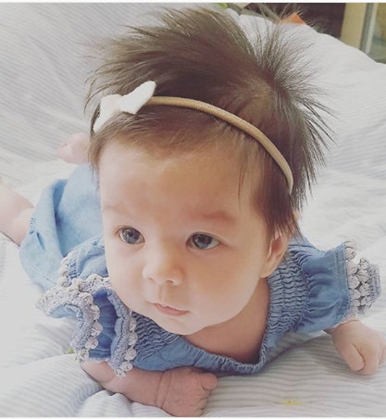 Op de afbeelding: Een babymeisje draagt een lichtblauwe denimjurk met witte bies en een wit strikbandje. Het bandje is een dun elastisch bandje met een klein wit strikje.