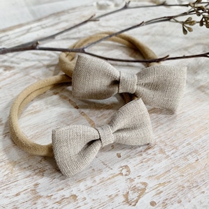 May include: Two beige fabric bow headbands. The bows are centered on the headbands and have a textured appearance. The headbands are a neutral color, and the bows are a slightly lighter shade. The background is a light wood surface.