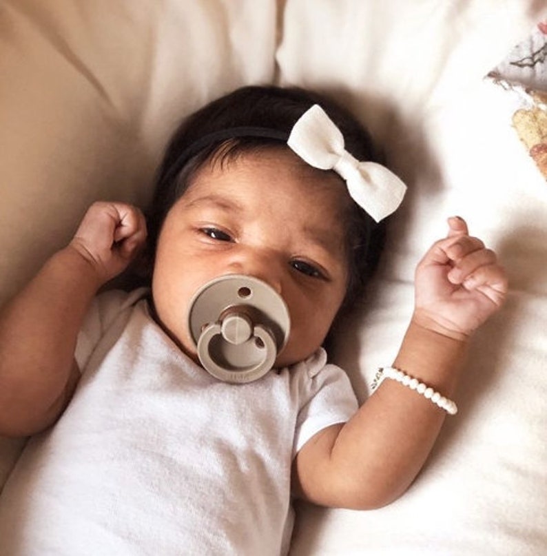 Op de afbeelding: Een baby die een wit rompertje draagt met een speen in de mond. De baby draagt een wit strikbandje en een witte kralen armband.