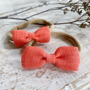 May include: Two coral-colored bow headbands. The bows have a textured appearance and are attached to neutral-toned elastic bands. The headbands are displayed on a white, weathered wooden surface, with a few sprigs of dried foliage in the background.