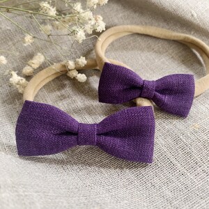 May include: Two purple bow headbands. The bows are made of a textured fabric and are attached to neutral-colored headbands. The bows are a deep purple color. The background is a neutral color.