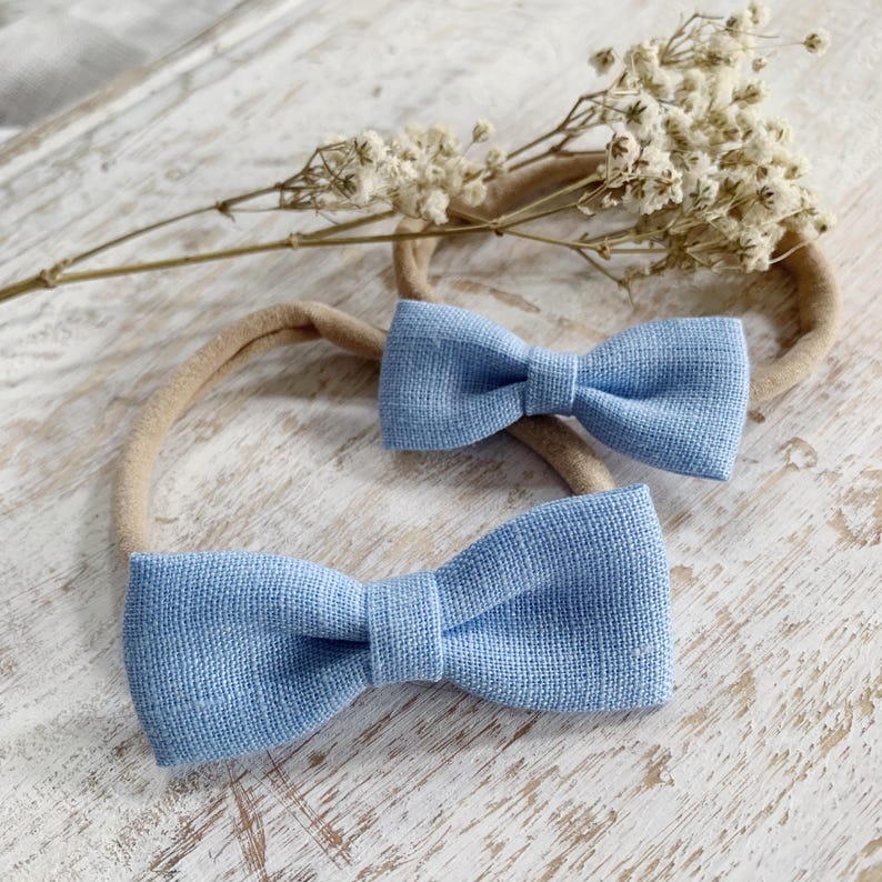 May include: Two light blue bow ties, one larger and one smaller, are displayed on a neutral-toned headband. The bows have a textured appearance. A sprig of dried white flowers is in the background, set against a light-colored wooden surface.