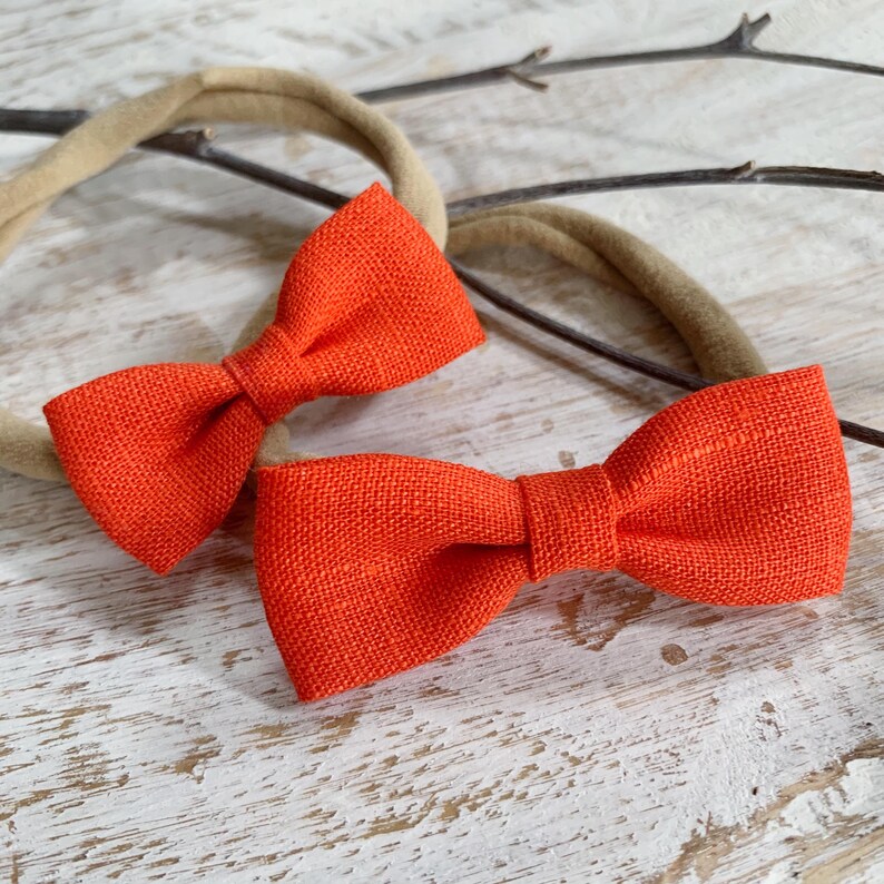 May include: Two bright orange bow headbands. The bows are made of a textured fabric and are attached to a neutral-colored headband. The bows are a vibrant shade of orange, and the headbands are suitable for babies and toddlers.
