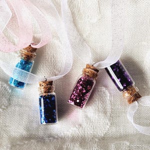 Pu&ograve; includere: Quattro piccole bottiglie di vetro con tappi di sughero, ciascuna riempita di glitter colorati. Le bottiglie sono sospese da nastri bianchi. I colori dei glitter includono blu, blu scuro, rosa e viola. Le bottiglie sono esposte su una superficie bianca strutturata.