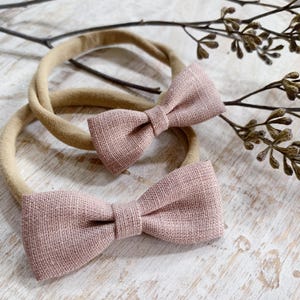 May include: Two baby headbands with dusty rose bowties. The bowties are made of a textured fabric and are attached to a neutral-colored elastic band. The headbands are displayed on a light-colored wooden surface with a sprig of dried foliage.