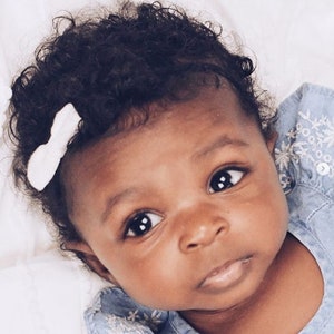Op de afbeelding: Een close-up van een baby met donker krullend haar, die een witte strik en een blauw-wit gestreept shirt draagt.