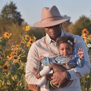 Op de afbeelding: Een man met een bruine fedora-hoed houdt een baby vast die een blauwe denim overall-jurk en een wit hoofdband draagt. De baby glimlacht en zwaait. De man en de baby staan in een veld met zonnebloemen.