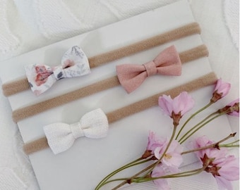 Puede incluir: Tres diademas elásticas de color nude con tres lazos de tela diferentes. Los lazos son un estampado floral blanco, un rosa claro sólido y un blanco sólido.
