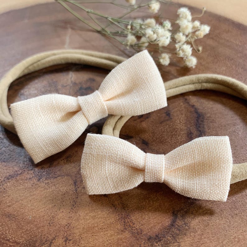 May include: Two peach-colored bow headbands are displayed on a wooden surface. The bows are made of textured fabric and are centered on beige elastic headbands. Dried flowers are visible in the background, adding a touch of natural decor.