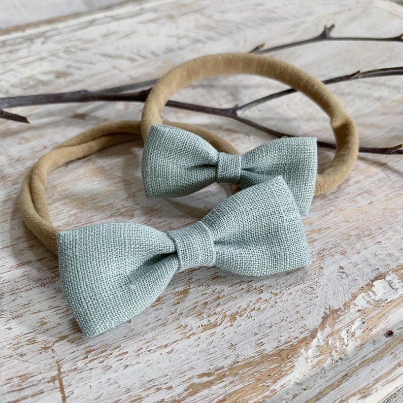 May include: Two sage green bow ties, each attached to a tan headband. The bows are made of a textured fabric and are tied in a classic bow shape. The headbands are circular and appear to be made of a soft, stretchy material. The background is a light-colored, weathered wooden surface.
