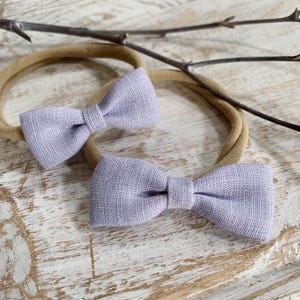 May include: Two light purple bow headbands. The bows are made of a textured fabric and are attached to a tan elastic band. The headbands are displayed on a light-colored wooden surface, with a few twigs in the background.