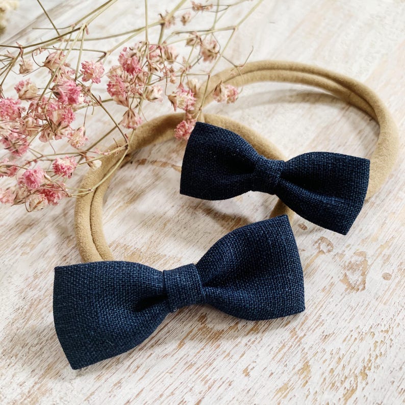 May include: Two navy blue bow ties on tan headbands. The bows are made of textured fabric and are centered on the headbands. The background includes dried pink flowers and a light-colored wooden surface.