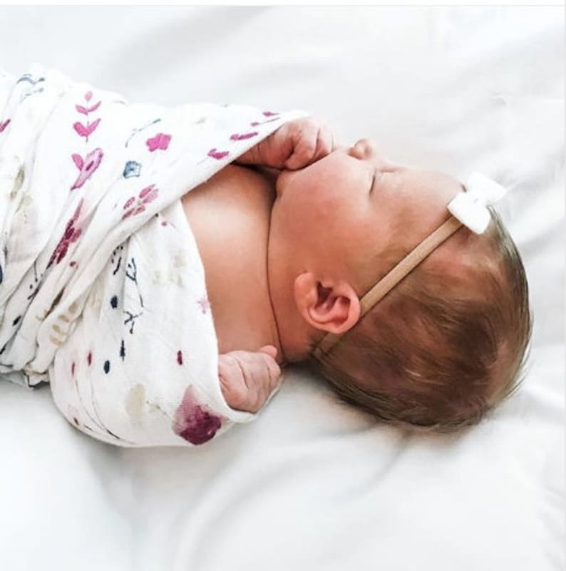 Op de afbeelding: Een pasgeboren baby gewikkeld in een witte swaddle met een roze en blauwe bloemenprint. De baby draagt ​​een wit strikbandje.