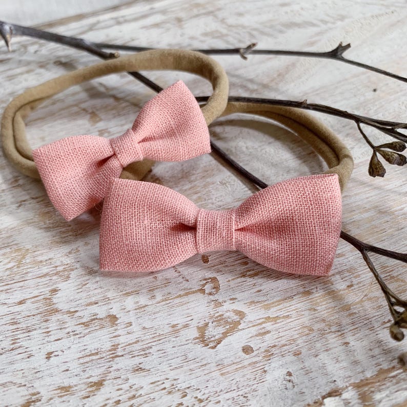 May include: Two pink linen bow headbands on a neutral-toned elastic band. The bows are a soft, muted pink and are centered on the elastic. The background is a light-colored, textured surface with a few small twigs.