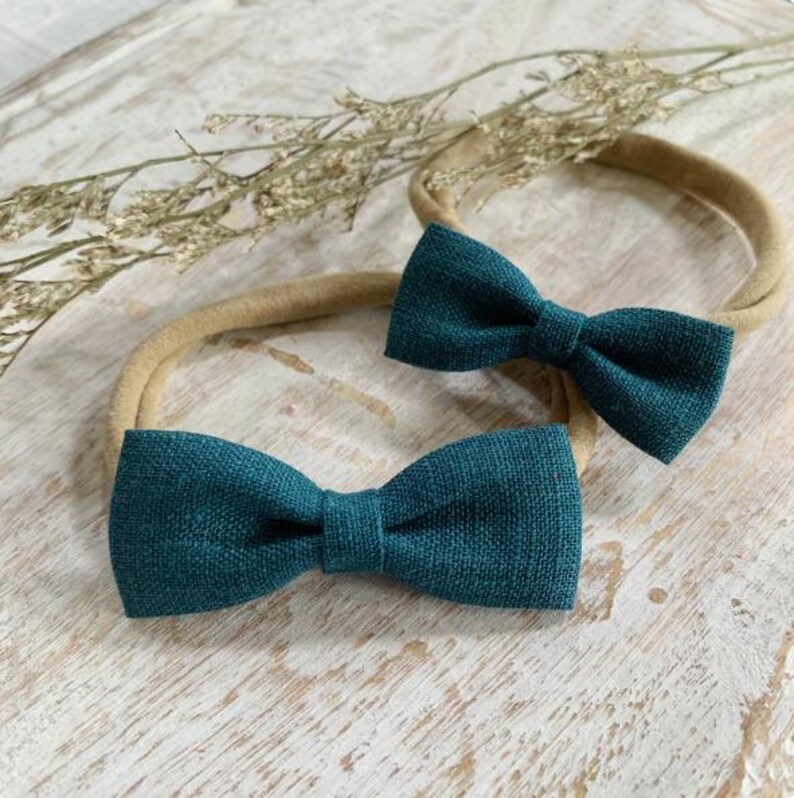 May include: Two teal-colored bow headbands are displayed on a light-colored wooden surface. The bows are made of textured fabric and are centered on beige elastic headbands. Dried floral accents are in the background.