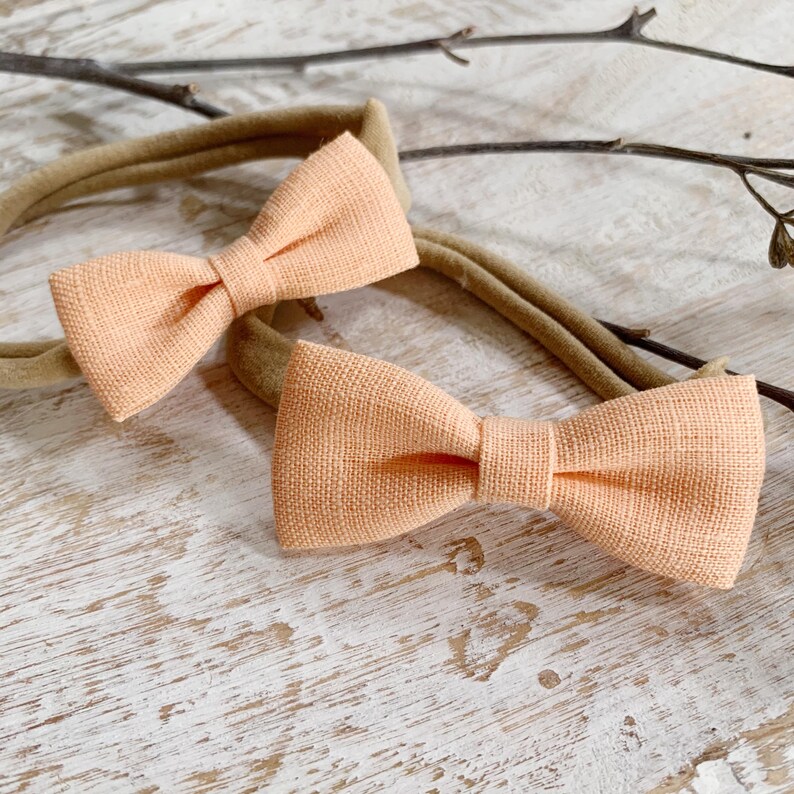May include: Two peach-colored bow ties with a textured fabric are displayed. Each bow is attached to a tan headband. The bows are centered in the frame, with a neutral background of wood and twigs.