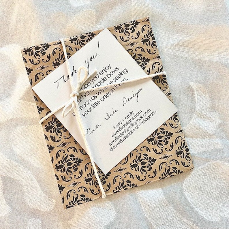Puede incluir: Un paquete envuelto en papel marr&oacute;n con un patr&oacute;n floral negro, atado con hilo. Una tarjeta blanca con texto escrito a mano, incluyendo "Thank you!" y el logo de Ever Iris Designs, est&aacute; adjunta.