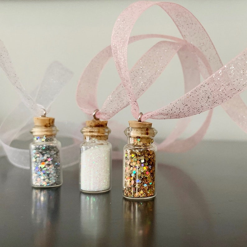 Pu&ograve; includere: Tre piccole bottiglie di vetro con tappi di sughero, riempite di glitter argento, bianco e oro. Ogni bottiglia &egrave; attaccata a un nastro scintillante bianco o rosa. Le bottiglie sono esposte su uno sfondo neutro.