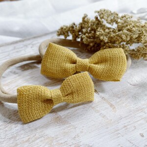 May include: Two mustard yellow bow headbands. The bows are made of a textured fabric and are attached to a neutral-colored headband. The bows are different sizes. Dried flowers are in the background.