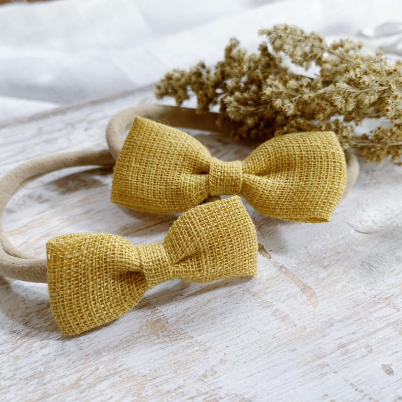May include: Two mustard yellow bow headbands. The bows are made of a textured fabric and are attached to a neutral-colored headband. The bows are different sizes. Dried flowers are in the background.