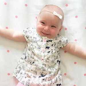 Op de afbeelding: Een baby draagt een wit shirt met korte mouwen met een zwart-wit dierenprint. De baby ligt op een wit laken met roze stippen en heeft een wit strikje in het haar.