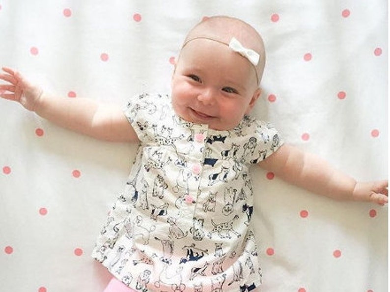 Op de afbeelding: Een baby draagt een wit shirt met korte mouwen met een zwart-wit dierenprint. De baby ligt op een wit laken met roze stippen en heeft een wit strikje in het haar.