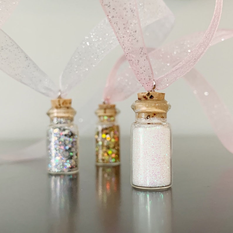 Pu&ograve; includere: Tre piccole bottiglie di vetro riempite di glitter argento, oro e bianco, ciascuna sormontata da un tappo di sughero e un nastro scintillante. Le bottiglie sono presentate su uno sfondo neutro, creando un effetto decorativo.