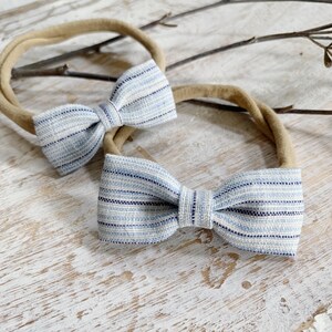 May include: Two baby headbands with light blue and navy striped bows. The bows are attached to tan elastic bands. The headbands are displayed on a light-colored, textured surface, with a few small twigs in the background.