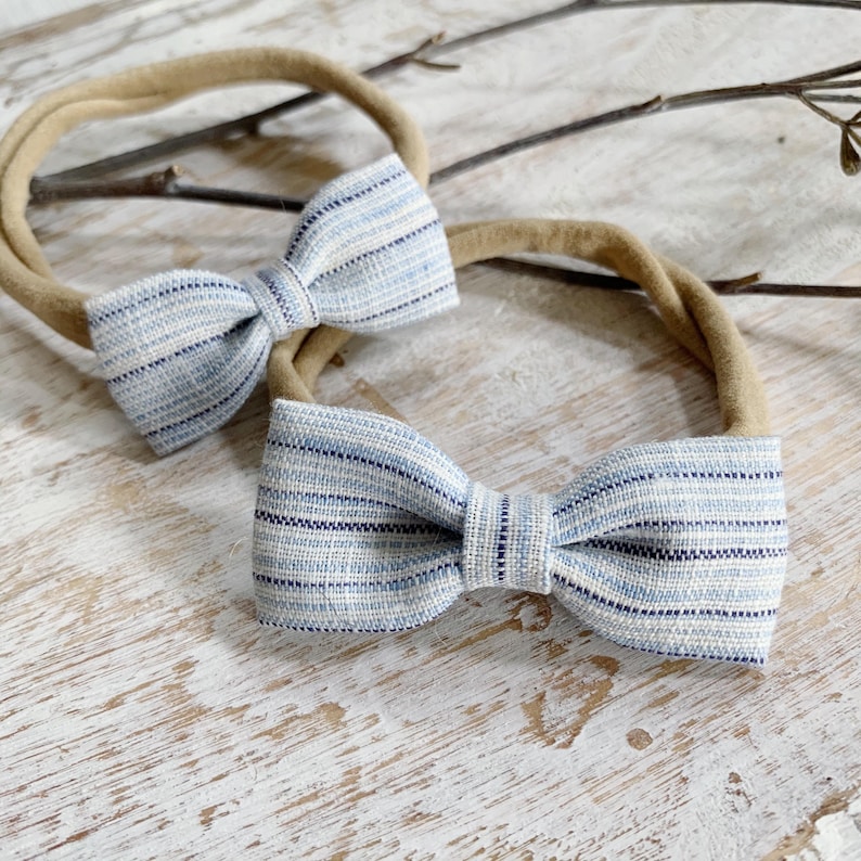 May include: Two baby headbands with light blue and navy striped bows. The bows are attached to tan elastic bands. The headbands are displayed on a light-colored, textured surface, with a few small twigs in the background.