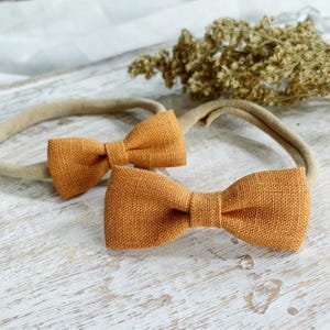 May include: Two orange-colored bow headbands. The bows are made of a textured fabric and are attached to a neutral-colored elastic band. The headbands are displayed on a white wooden surface with dried floral accents in the background.