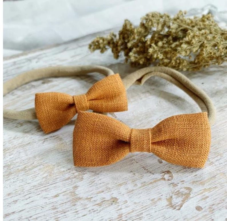 May include: Two orange-colored bow headbands. The bows are made of a textured fabric and are attached to a neutral-colored elastic band. The headbands are displayed on a white wooden surface with dried floral accents in the background.