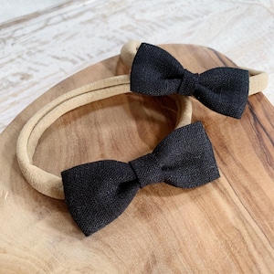 May include: Two black bow headbands with a textured fabric are displayed on a wooden surface. The headbands have a neutral-colored band. The bows are centered on the band.