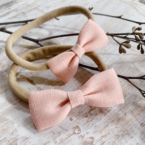 May include: Two baby headbands with pink bowties. The bowties are made of a textured fabric and are attached to a neutral-colored elastic band. The headbands are displayed on a light-colored wooden surface with decorative twigs.