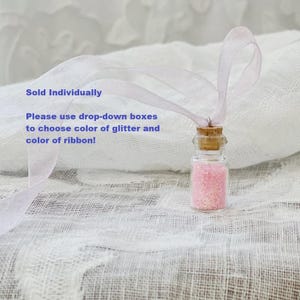 Pu&ograve; includere: Una piccola bottiglia di vetro piena di glitter rosa, sigillata con un tappo di sughero e legata con un nastro rosa chiaro. Il testo nell'immagine recita: "Venduto singolarmente. Si prega di utilizzare i menu a tendina per scegliere il colore dei glitter e del nastro!"