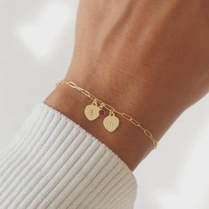 May include: A gold bracelet with a delicate chain. It features two heart-shaped charms and a small red gemstone. One heart charm has the letter "A" engraved, and the other has the letter "M".