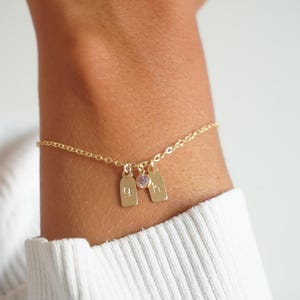 Peut inclure: Un bracelet doré délicat avec deux petits charms rectangulaires et une petite pierre précieuse violette. Les charms portent les lettres "g" et "h". Le bracelet est porté au poignet.