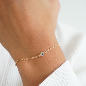May include: A fine gold bracelet featuring a small, round, pale blue gemstone. The bracelet is worn on a wrist, with the gold chain clearly visible. The person is wearing a white long-sleeved top.