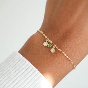 Peut inclure: Un bracelet en or délicat avec trois breloques. Deux breloques rondes avec les lettres "A" et "M" et une petite breloque en forme de cœur vert. Le bracelet est sur un poignet, et le fond est blanc.