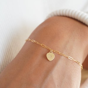 May include: A gold chain bracelet with a heart charm featuring the letter "M" engraved on it.