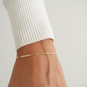 May include: A delicate gold chain bracelet featuring a flat mariner link design. The bracelet is worn around the wrist, showcasing a polished metallic finish that catches the light. The links are uniform in size and shape, creating a continuous, refined look.